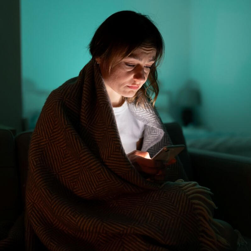 Person searching for therapist on their smartphone in the dark wrapped in blanket with worried look on face